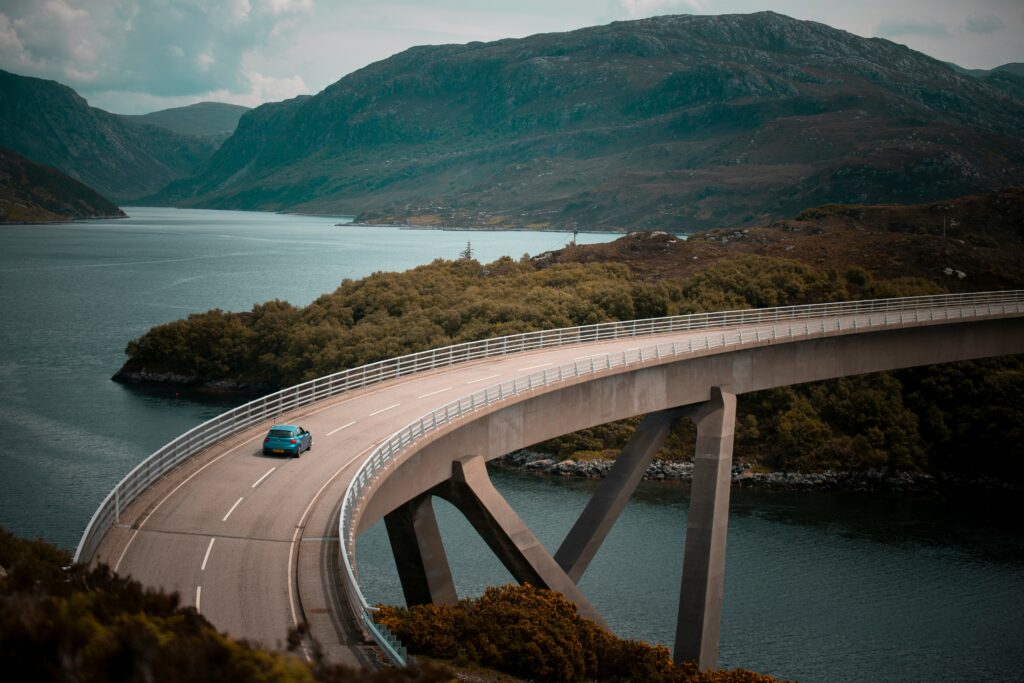 Auto, das eine nach rechts gekrümmte Brücke entlangfährt. Im Hintergrund schöne Berglandschaft, die von einem Fluss geteilt wird